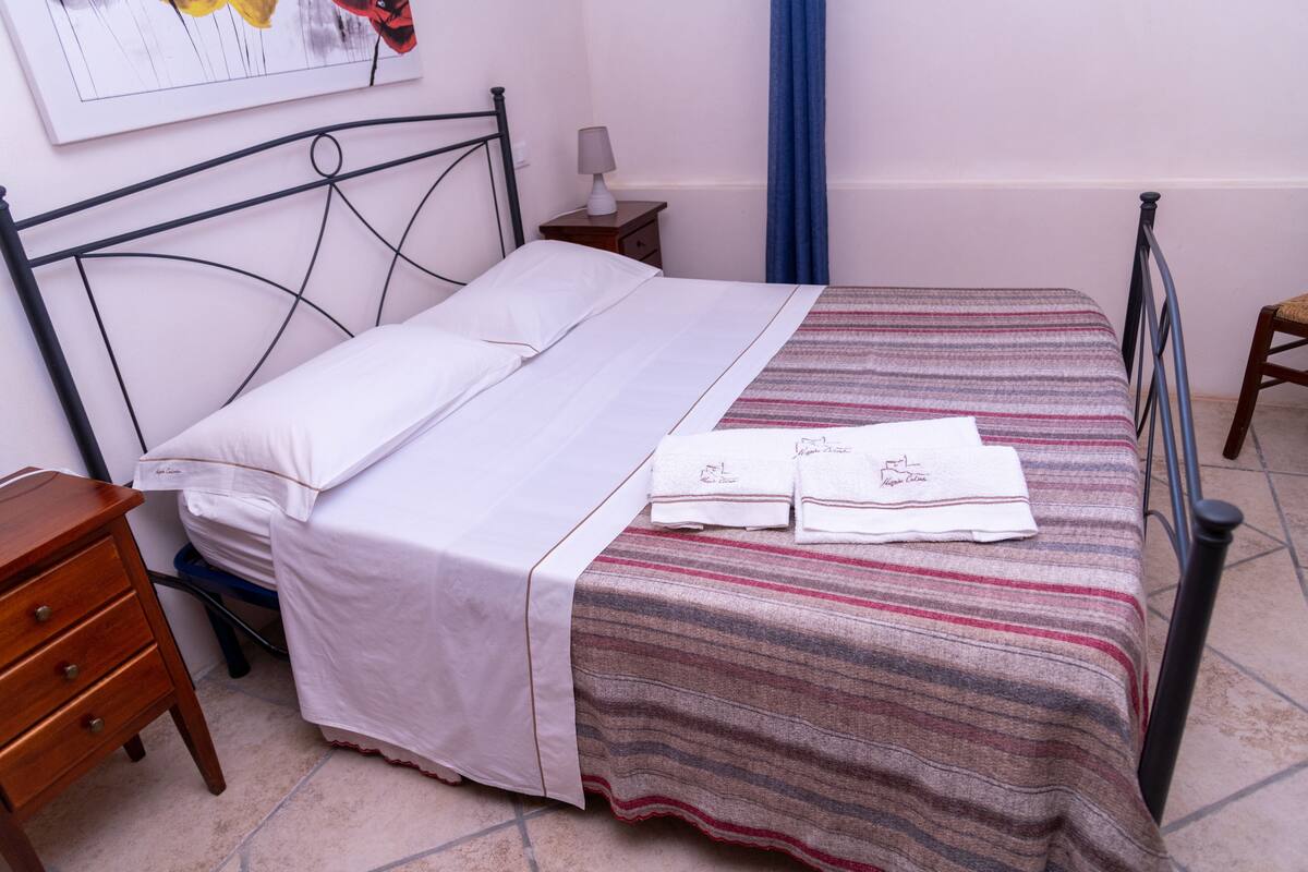 A neatly made bed with a striped bedspread and two towels, a wooden nightstand with a lamp, and a framed picture on the wall.