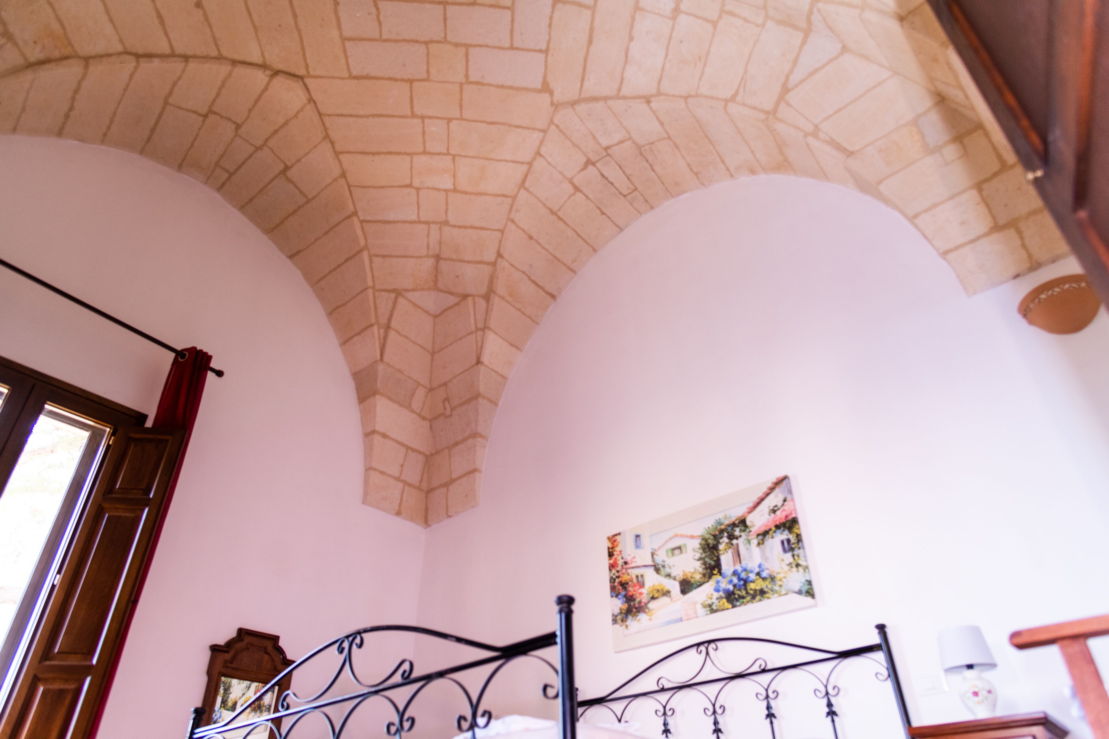 A room with a brick ceiling, a bed with a metal frame, a wooden door, and a wall-mounted picture.