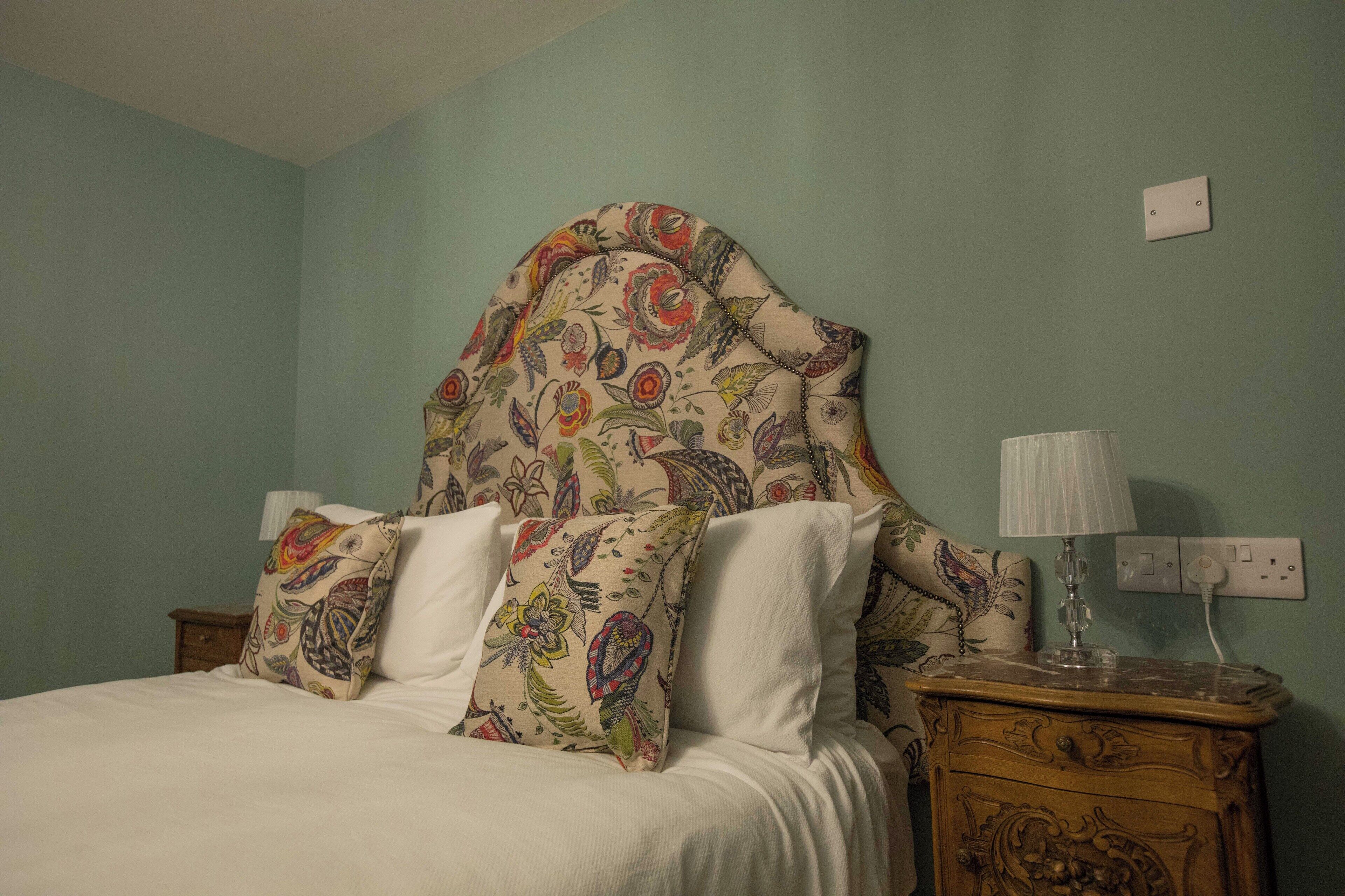 A bed with floral-patterned pillows, a wooden nightstand with a lamp, and a wall socket.