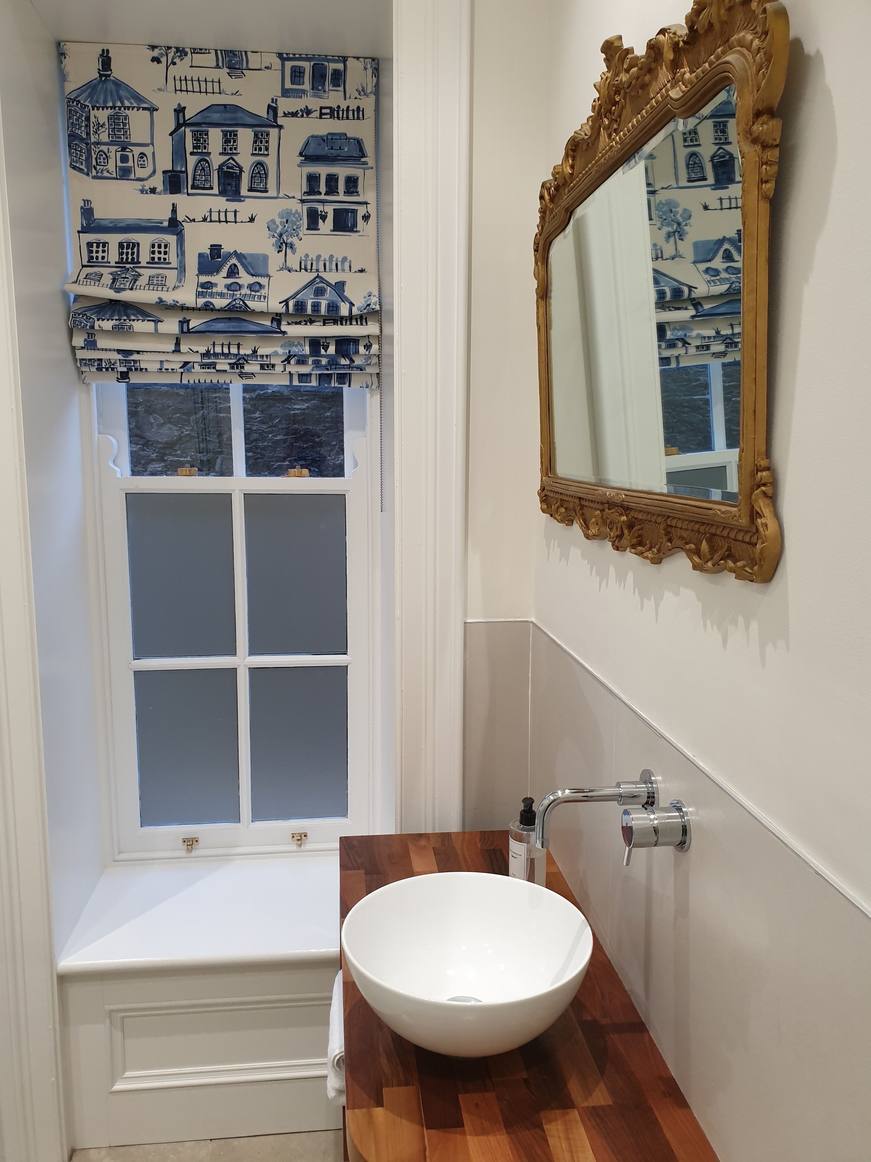 Un baño con un lavamanos blanco, un tocador de madera, un espejo con marco dorado y una ventana con un cortinero azul y blanco.