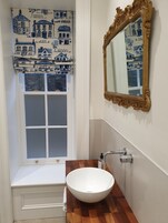 Un baño con un lavamanos blanco, un tocador de madera, un espejo con marco dorado y una ventana con un cortinero azul y blanco.