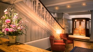 A grand staircase with ornate railings, a red armchair, a table with a lamp, and a large floral arrangement. - Bishop's Gate Hotel (Londonderry)