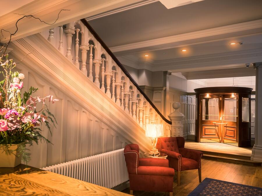 A grand staircase with ornate railings, a red armchair, a table with a lamp, and a large floral arrangement.
