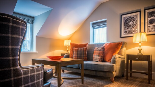 A cozy living room with a plaid armchair, a sofa with plaid cushions, a wooden table with a bowl, two lamps, and framed pictures on the wall. - Bishop's Gate Hotel (Londonderry)
