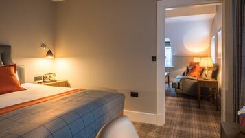 A hotel room with a bed, a nightstand, a lamp, a chair, and a view through an open door to another room with a sofa and a lamp.