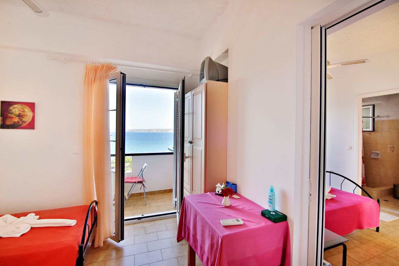 Una habitación con cama, una mesa con mantel rosa, una silla y vista al mar por una puerta abierta.