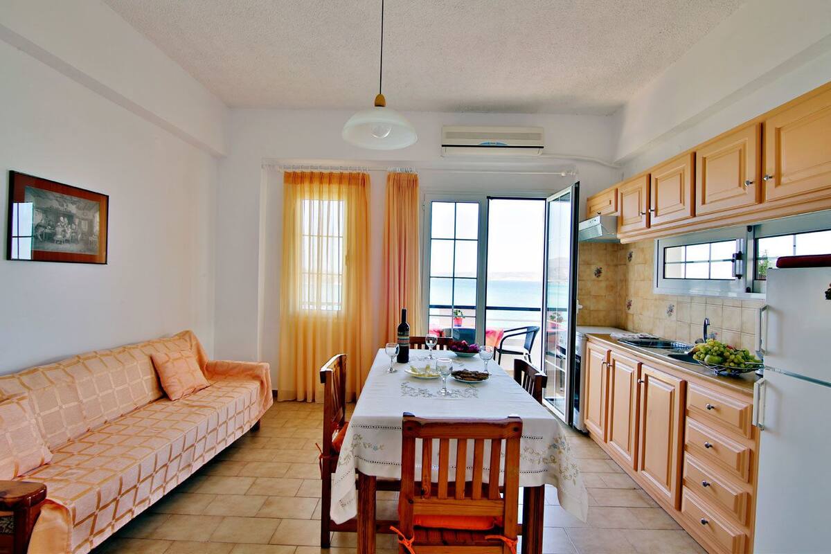 Una sala de estar acogedora con sofá, mesa de comedor y sillas, una cocina con armarios de madera, y un gran ventanal con vistas al mar.