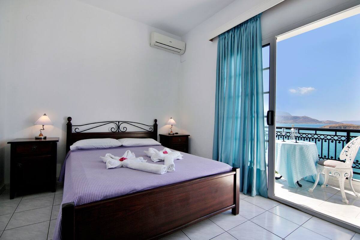 Habitación de hotel con cama, dos mesitas de noche, ventana con cortinas y balcón con mesa y silla.