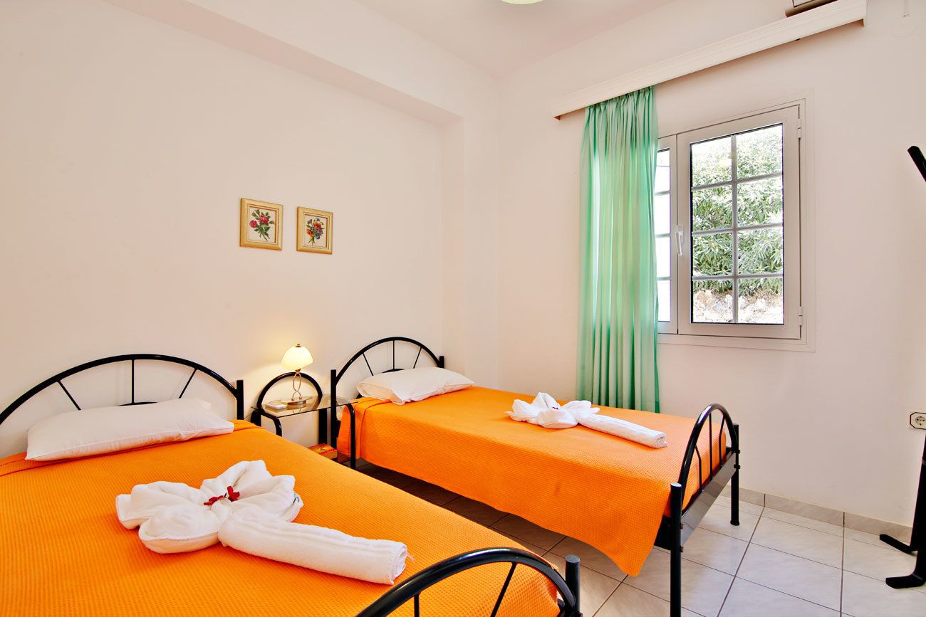 Une chambre avec deux lits, chacun recouvert d’une couvre-lit orange et agrémenté d’oreillers blancs. Deux tableaux encadrés ornent le mur, une fenêtre est habillée de rideaux verts, et une table de chevet porte une lampe.