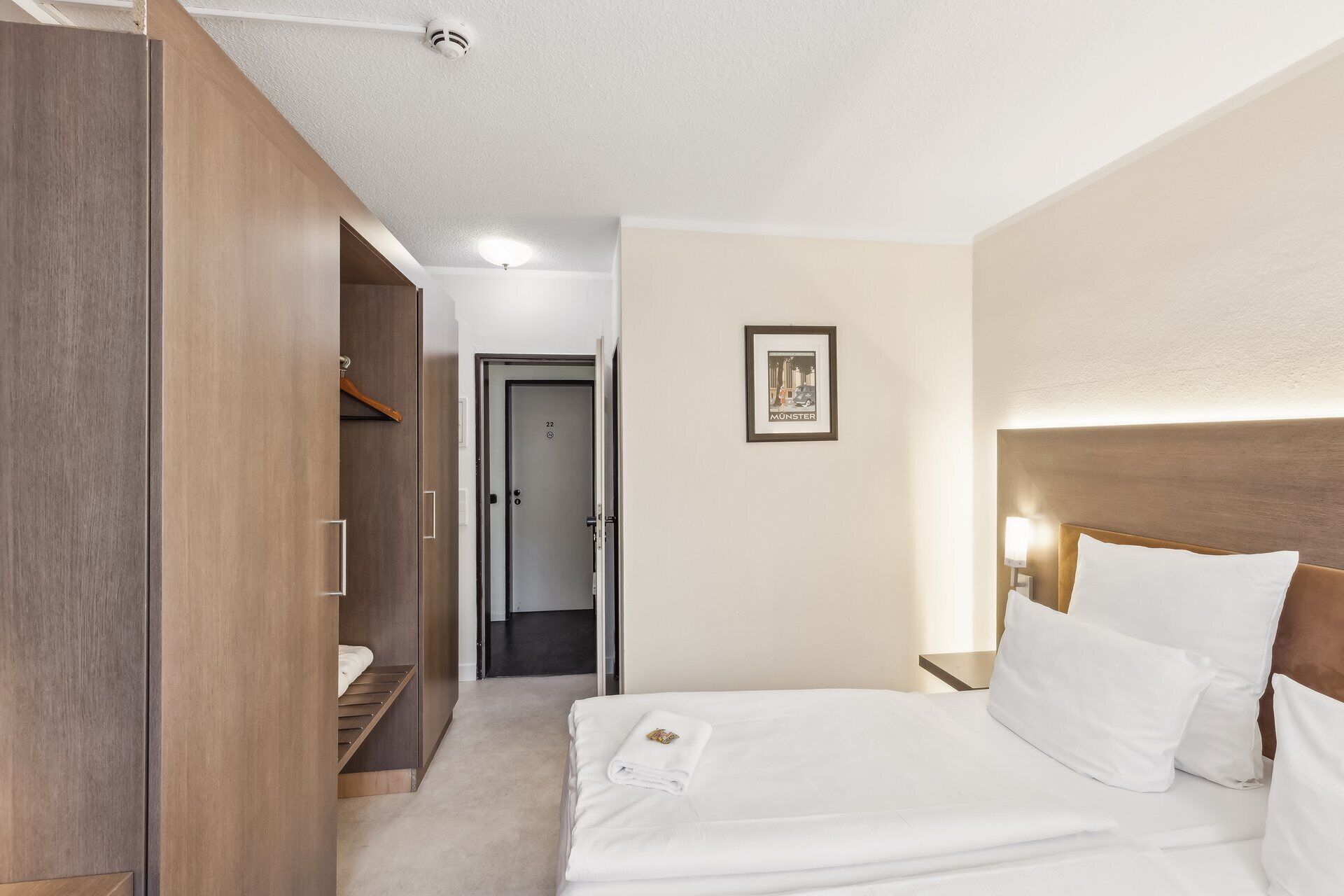 A hotel room with a bed, a wardrobe, a door, a framed picture on the wall, and a light fixture above the bed.