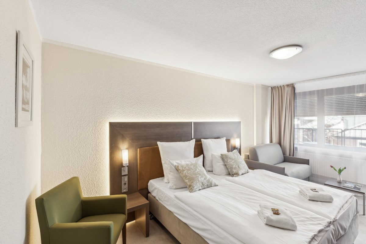 A hotel room with a large bed, a green armchair, a bedside table with a lamp, and a window with curtains.
