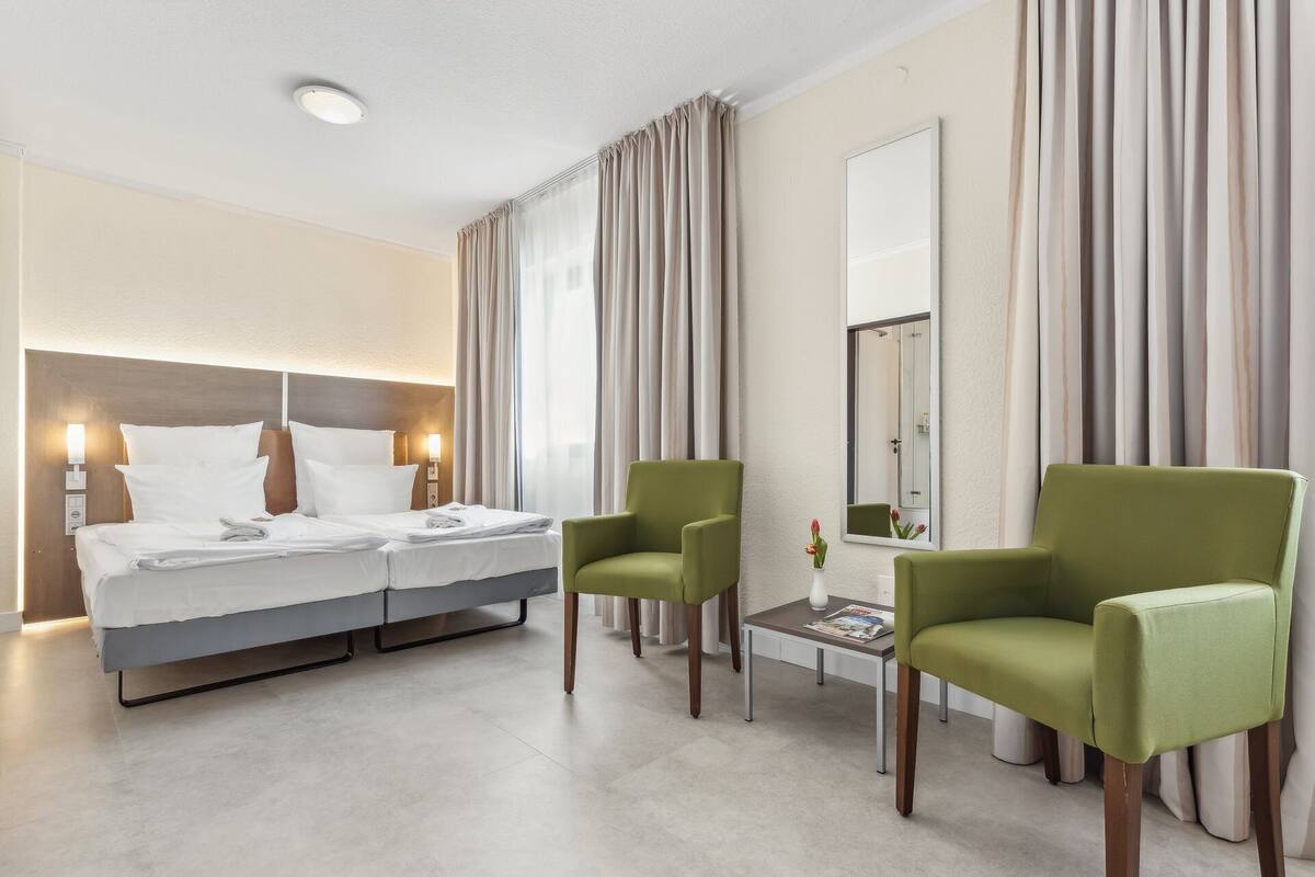 A hotel room with a bed, two chairs, a small table with a magazine, a mirror, and curtains.