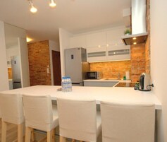 A modern kitchen with white cabinets, a brick accent wall, a stainless steel refrigerator, a microwave, and a kettle. There are four white chairs around a table with a water bottle on it.
