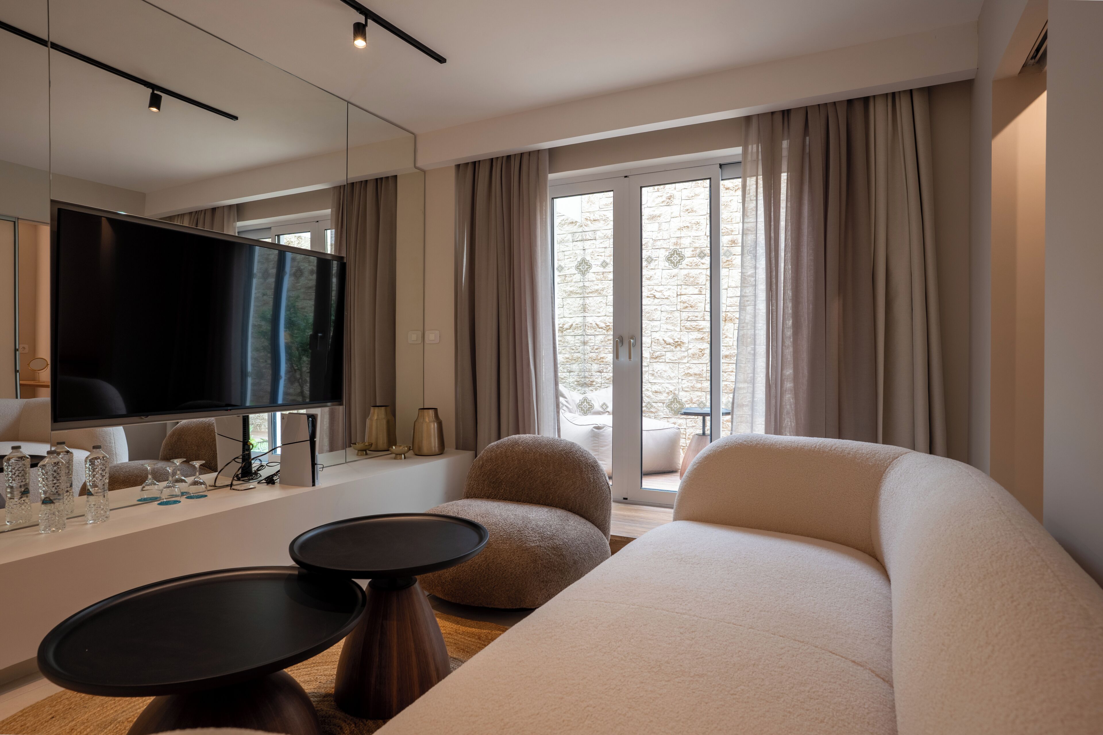 A modern hotel room with a large flat-screen TV, comfortable seating, a balcony with curtains, and a view of the outdoors.