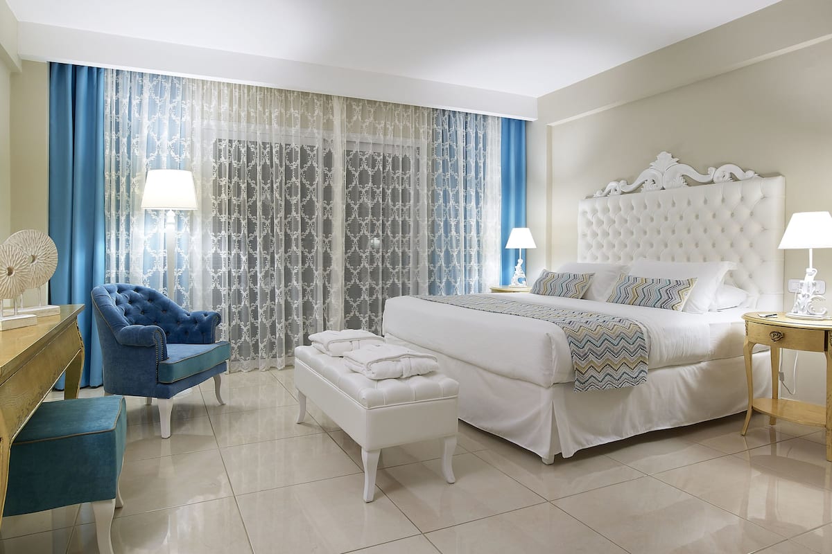 Una habitación de hotel con una cama grande, un sillón azul, una mesita pequeña y un ventanal con cortinas traslúcidas.