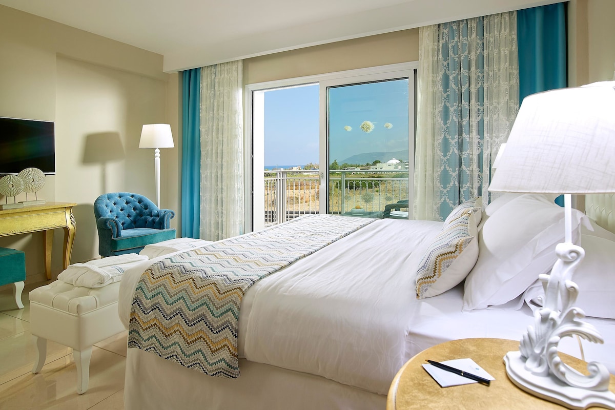 Habitación de hotel con cama, escritorio, silla, televisor y balcón con vistas.