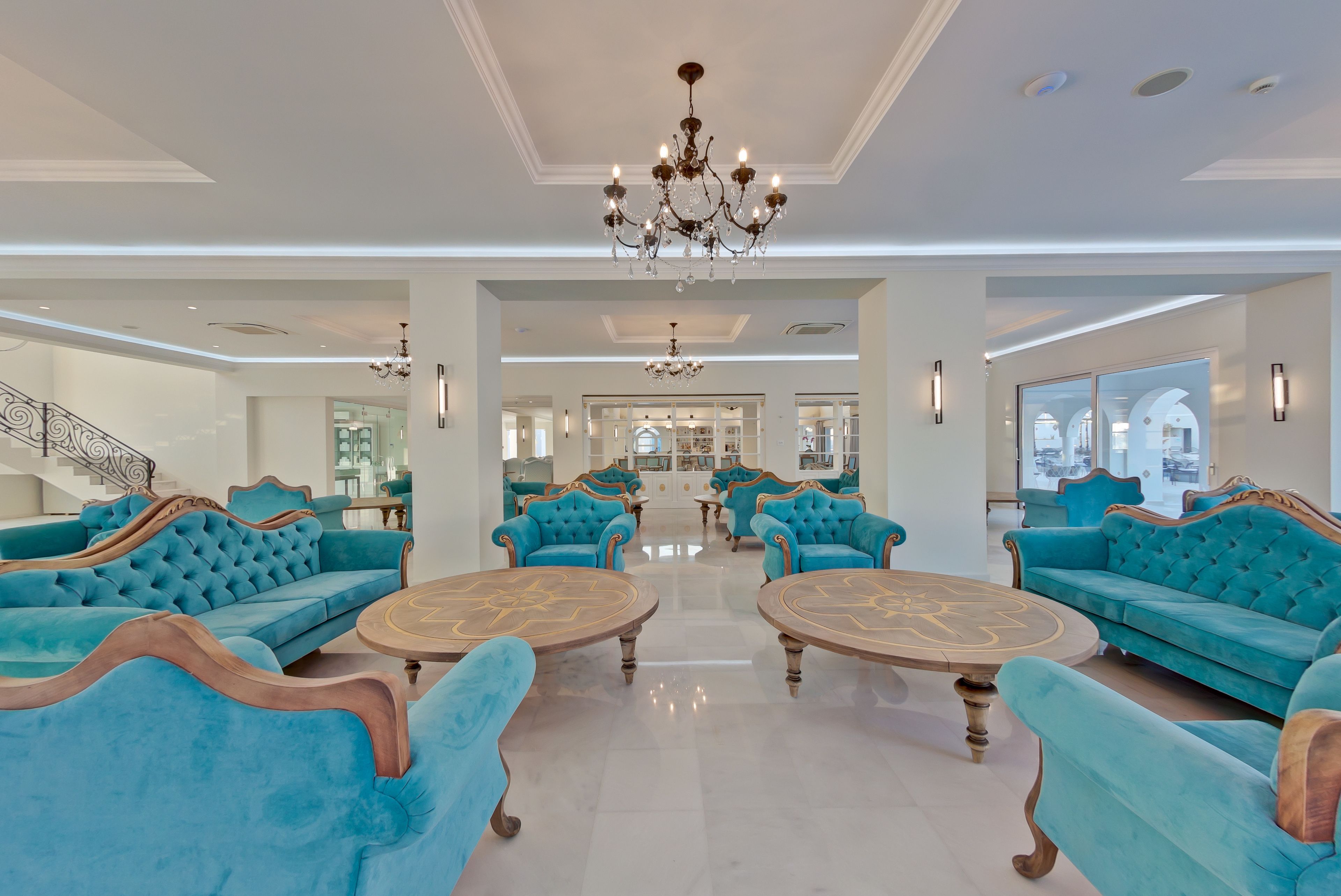 A spacious living room with teal sofas and round coffee tables, a grand chandelier, and a modern staircase.