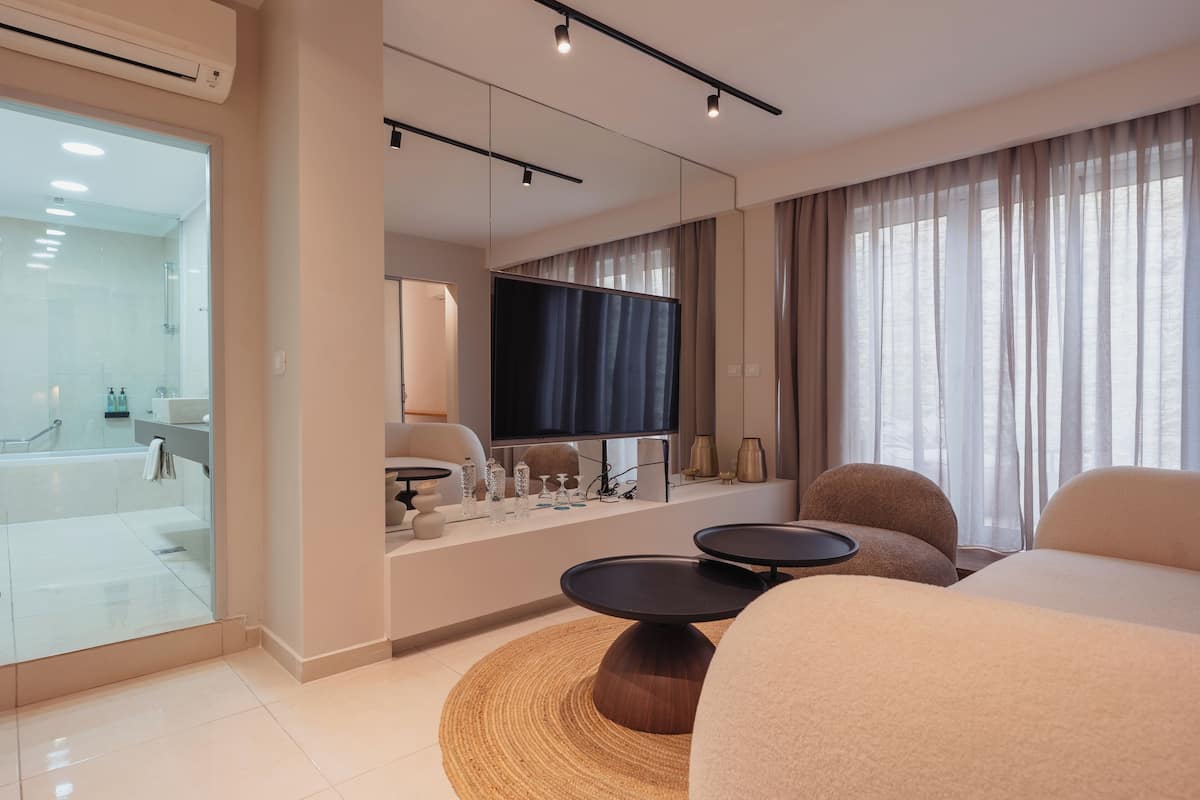 Una habitación de hotel moderna con una cama grande, un televisor, un área de estar con una mesa y un área de ducha con paredes de vidrio.