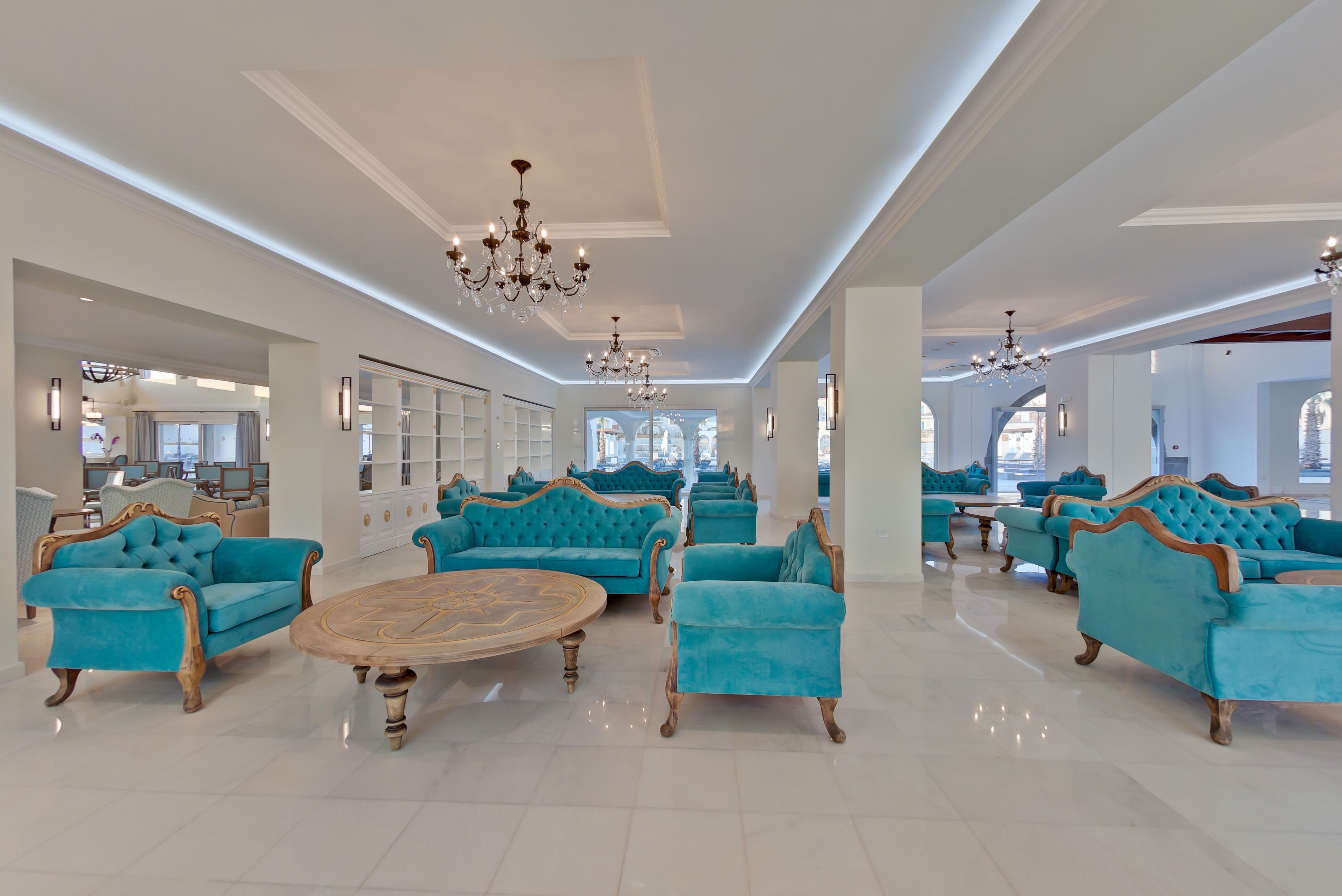 A spacious lobby with teal upholstered sofas arranged around a central wooden coffee table, large windows providing natural light, and a chandelier hanging from the ceiling.