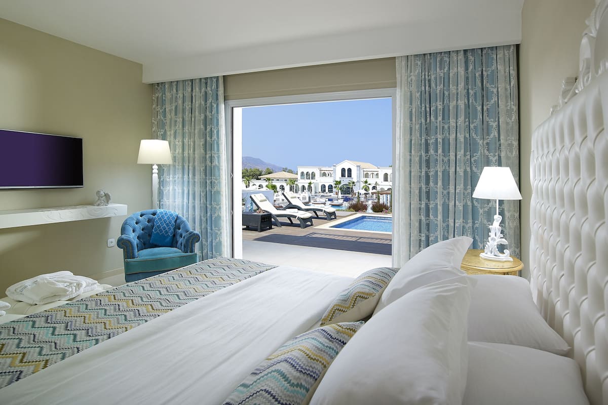 Habitación de hotel con cama, televisor, sillón y vistas a una piscina y edificios.