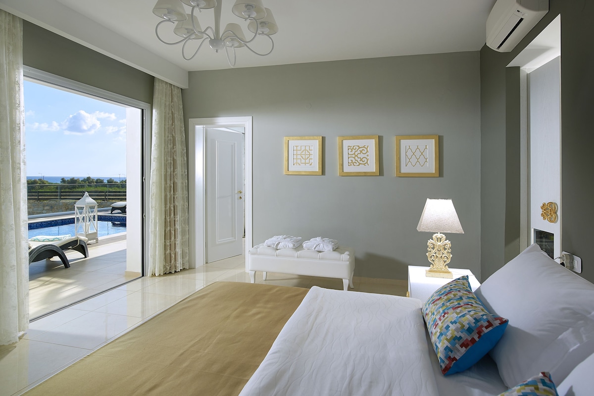 A hotel room with a bed, a nightstand, a lamp, and a view of the sea through a large window.