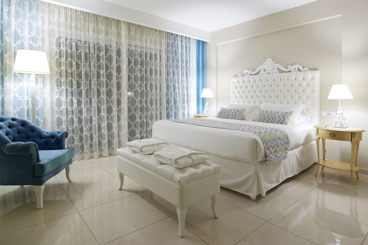 Una habitación de hotel con una cama grande, un sillón azul, un reposapiés blanco y una mesita de noche con una lámpara.