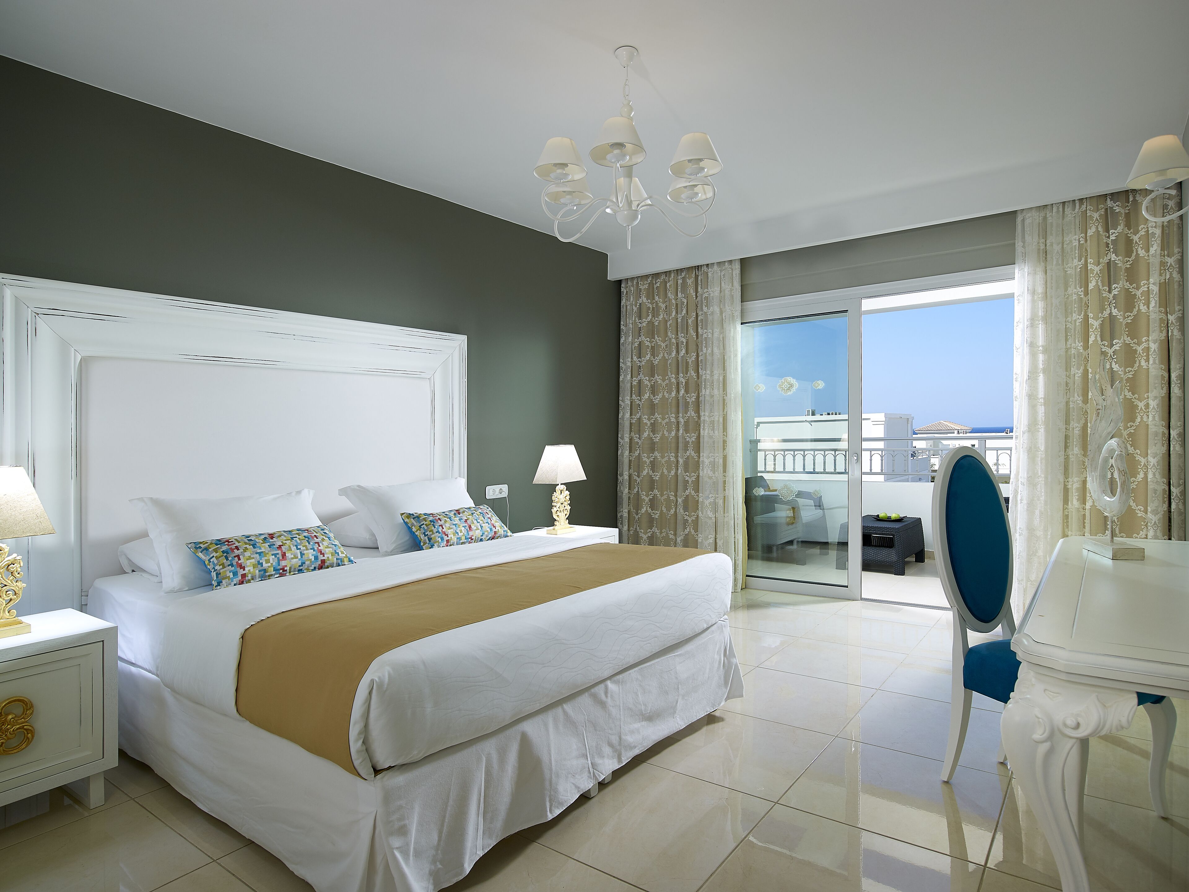 A hotel room with a large bed, a nightstand with a lamp, a chair, and a view of the sea through a balcony.