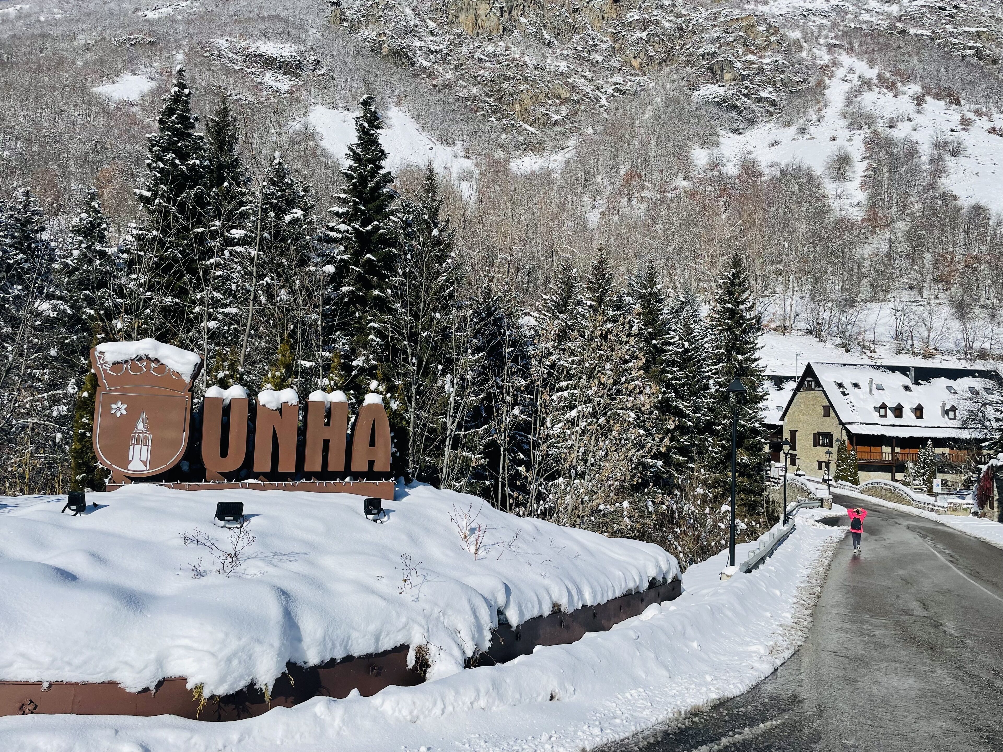 Un letrero cubierto de nieve con la palabra 'UNHA' y un escudo, rodeado de árboles cargados de nieve y un paisaje montañoso.