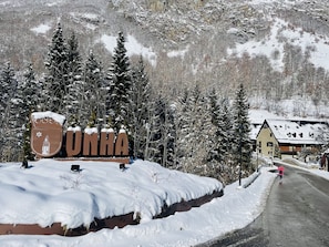 Un letrero cubierto de nieve con la palabra 'UNHA' y un escudo, rodeado de árboles cargados de nieve y un paisaje montañoso.