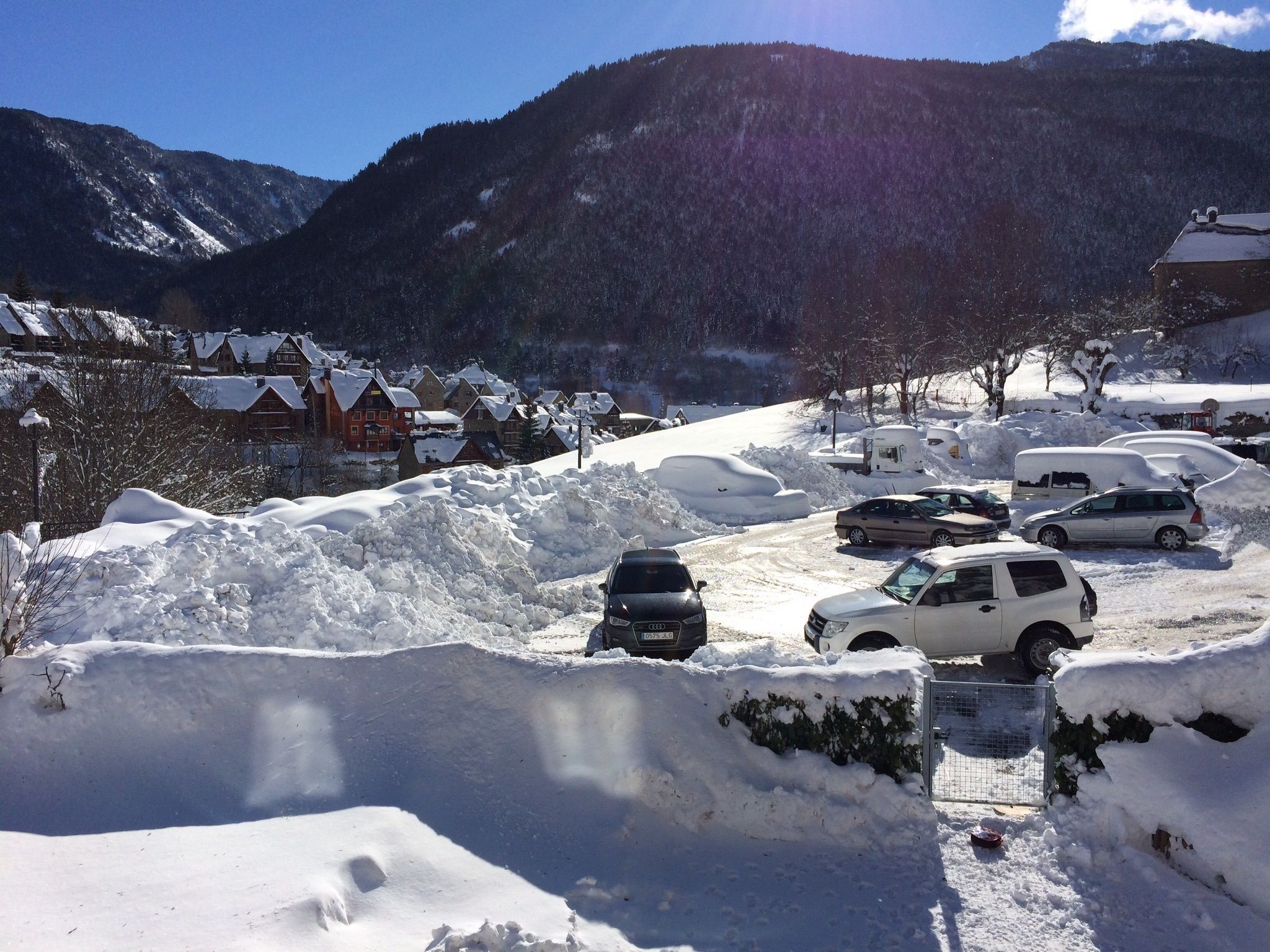 A snowy parking lot with several cars and a snowbank in the foreground.