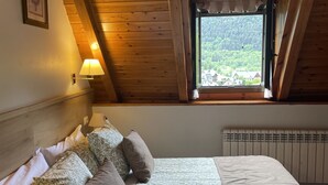 Une chambre confortable avec un plafond en bois, une fenêtre donnant sur une forêt et un lit agrémenté d’une literie à motifs floraux.
