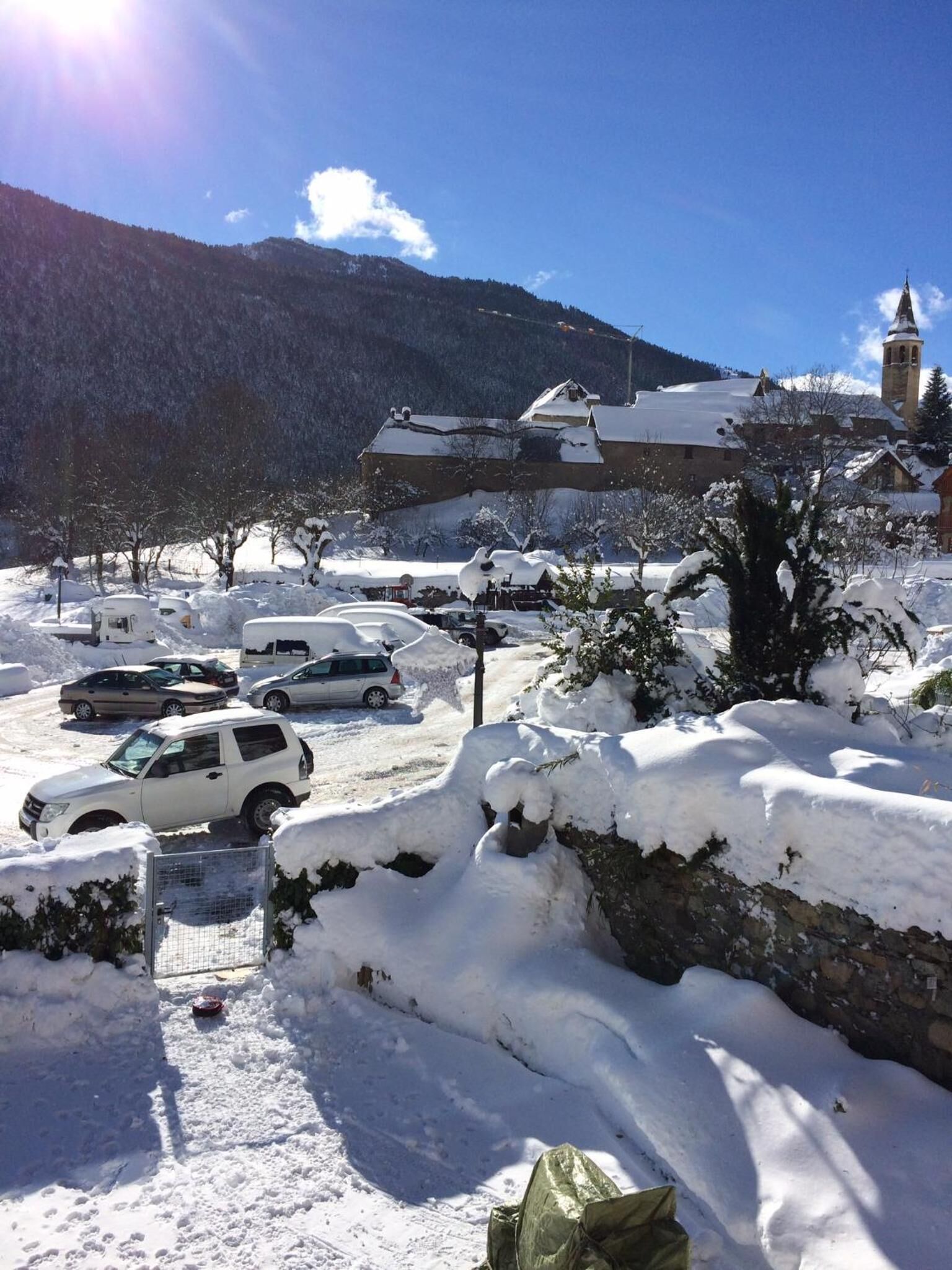 A snowy parking area with several cars, a stone wall, and a building with a clock tower in the background.