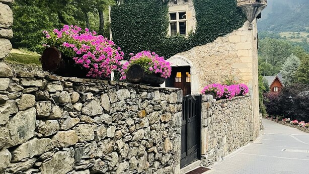 Un bâtiment en pierre doté d’une tourelle et de murs couverts de lierre, un chemin pavé et des fleurs roses.