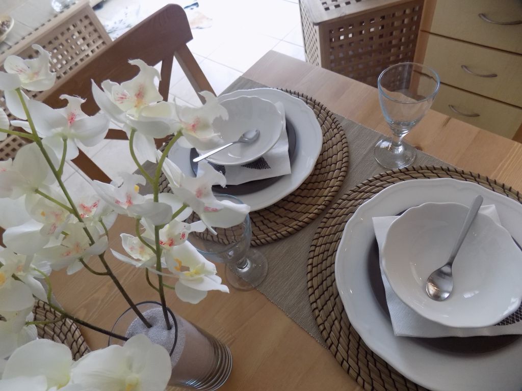 Una mesa de comedor dispuesta con platos blancos, cubiertos y un vaso de agua, rodeada de individuales tejidos y un jarrón de orquídeas blancas.