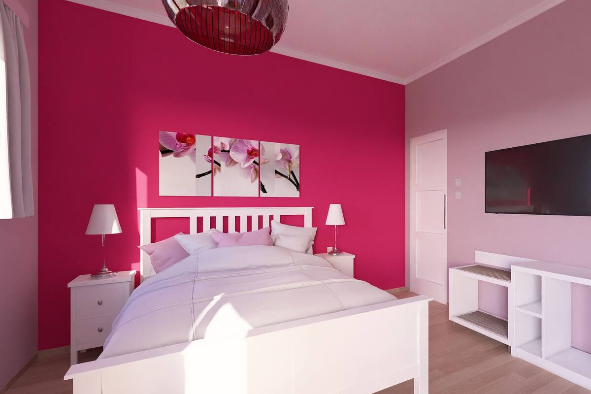 Un dormitorio con una cama blanca, paredes rosas, una lámpara en el techo, un televisor y una mesita de noche blanca.