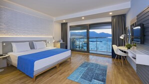 Een moderne hotelkamer met een groot bed, een bureau met stoel, een televisie en een balkon met uitzicht op zee en bergen.