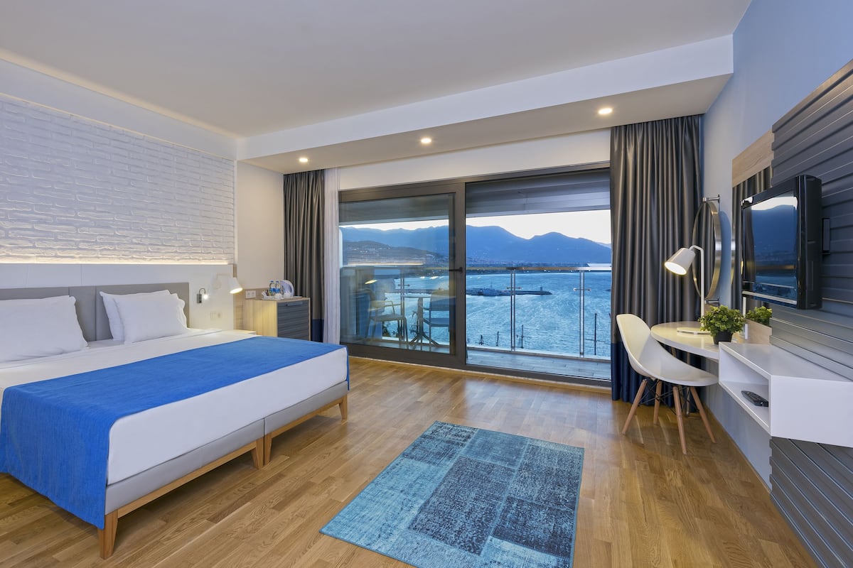 A modern hotel room with a large bed, a desk with a chair, a TV, and a balcony with a view of the sea and mountains.