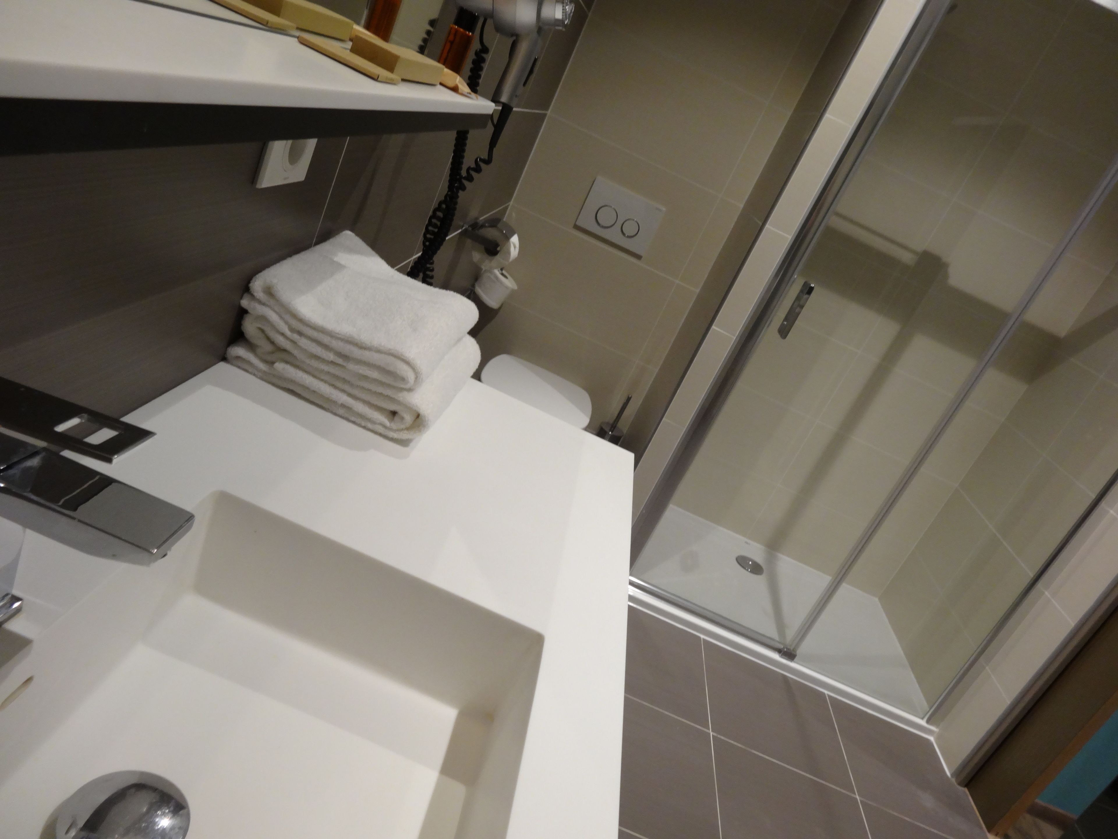 A modern bathroom with a white sink, a glass shower enclosure, and a towel stack.
