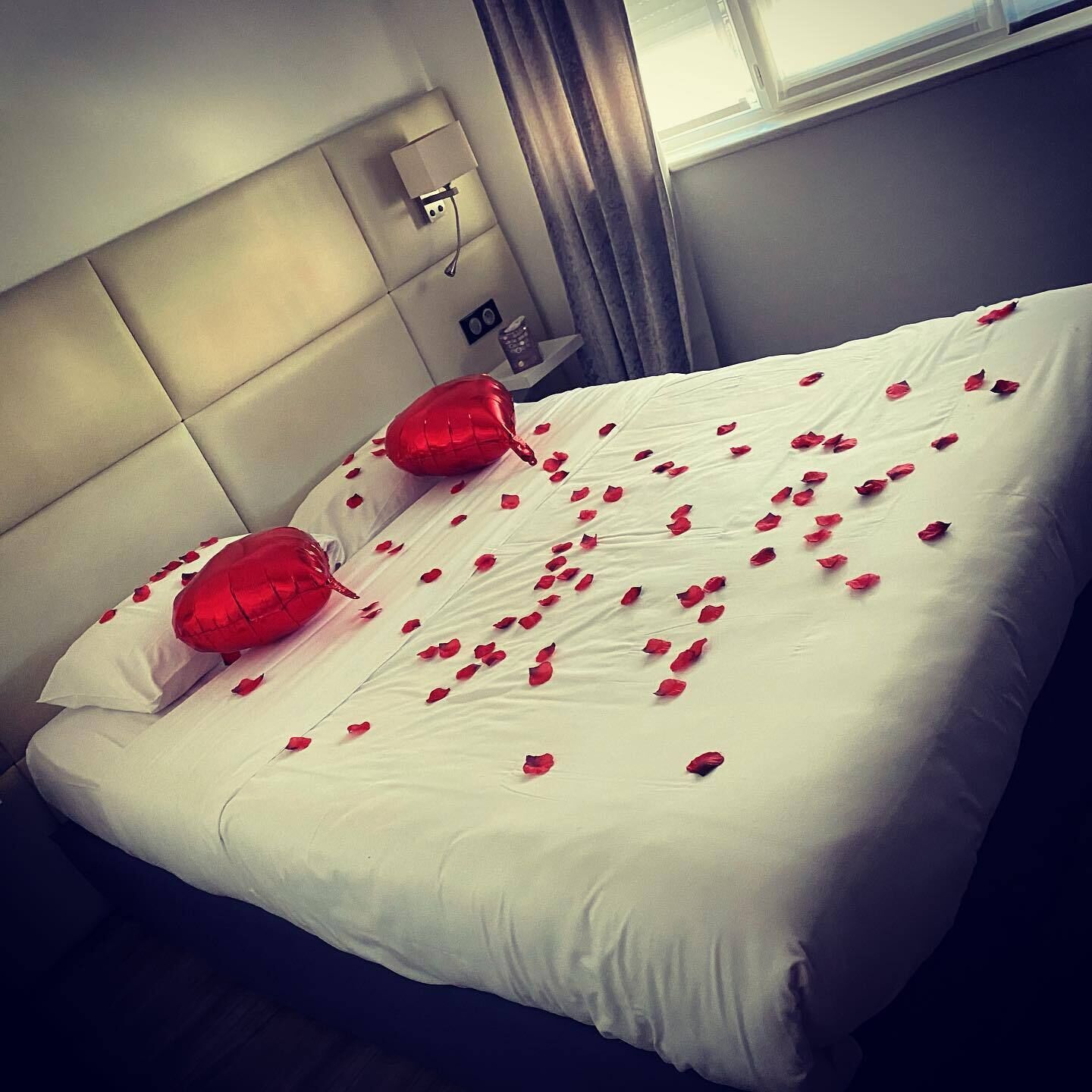 Una cama bien hecha con ropa blanca y pétalos de rosa roja esparcidos.