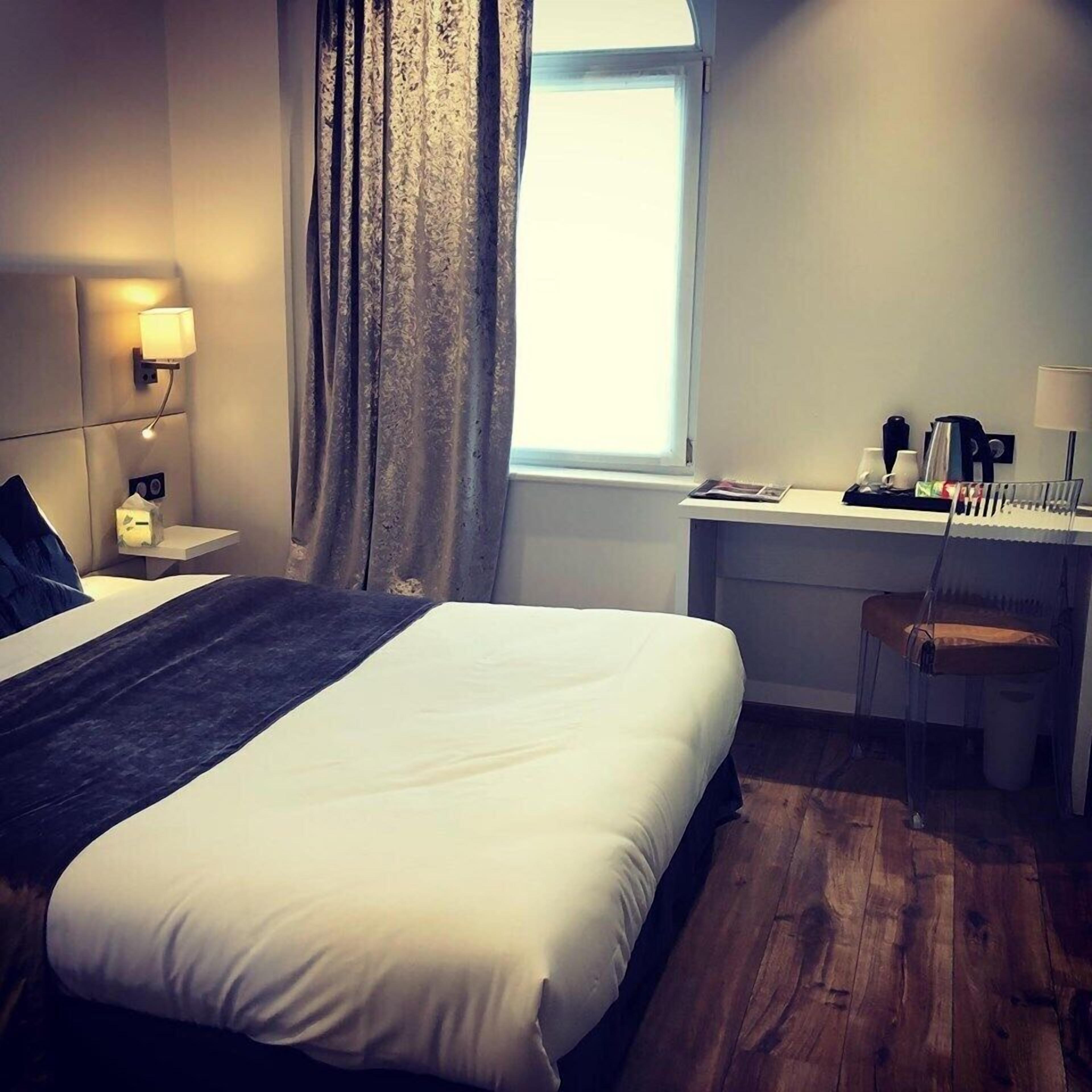A hotel room with a large bed, a desk, a chair, a window with curtains, and a wall-mounted lamp.