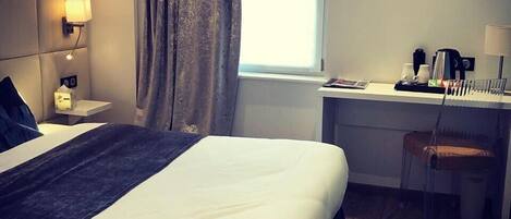 Habitación de hotel con una cama grande, un escritorio, una silla, una ventana con cortinas y una lámpara de pared.