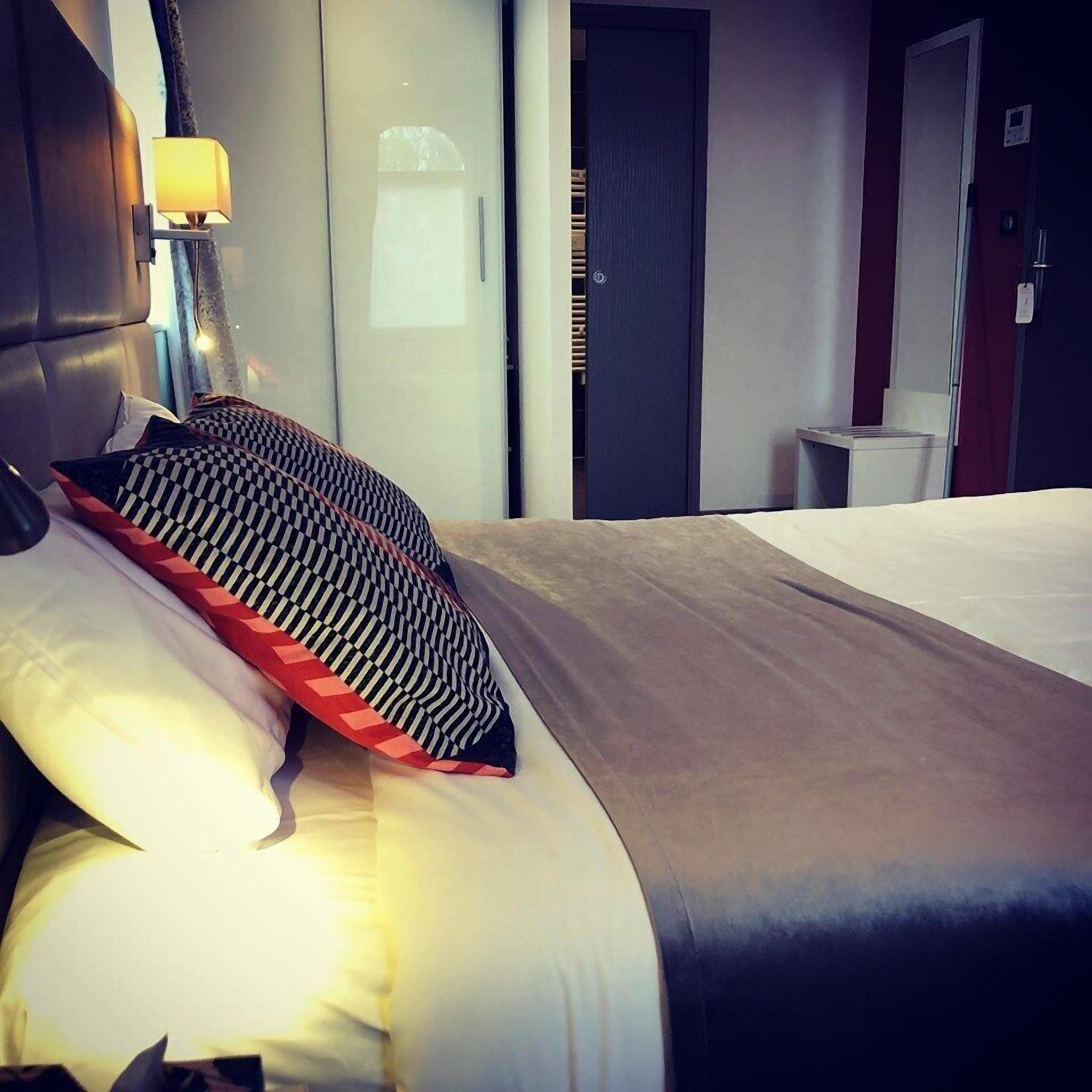 A neatly made bed with a headboard, a bedside lamp, and a view of a hallway with a door.