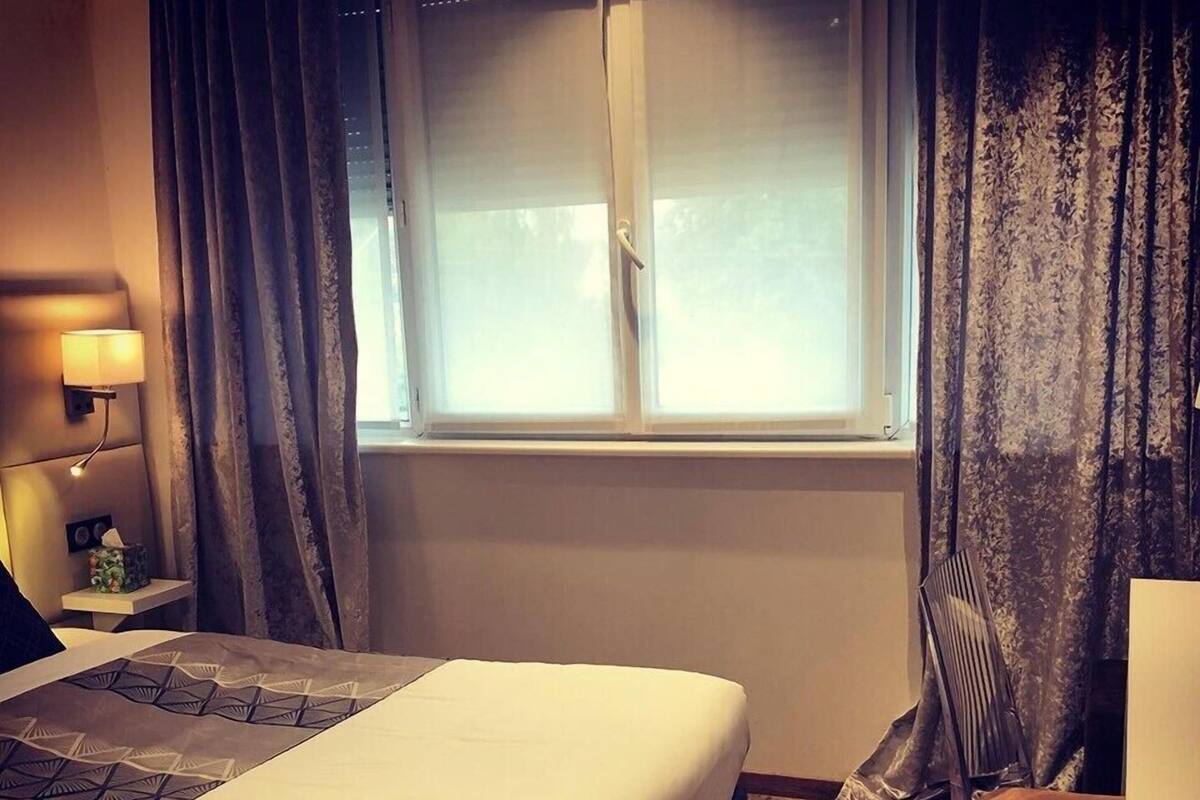 Una habitación de hotel con cama, mesita de noche, lámpara, silla y ventana con cortinas.