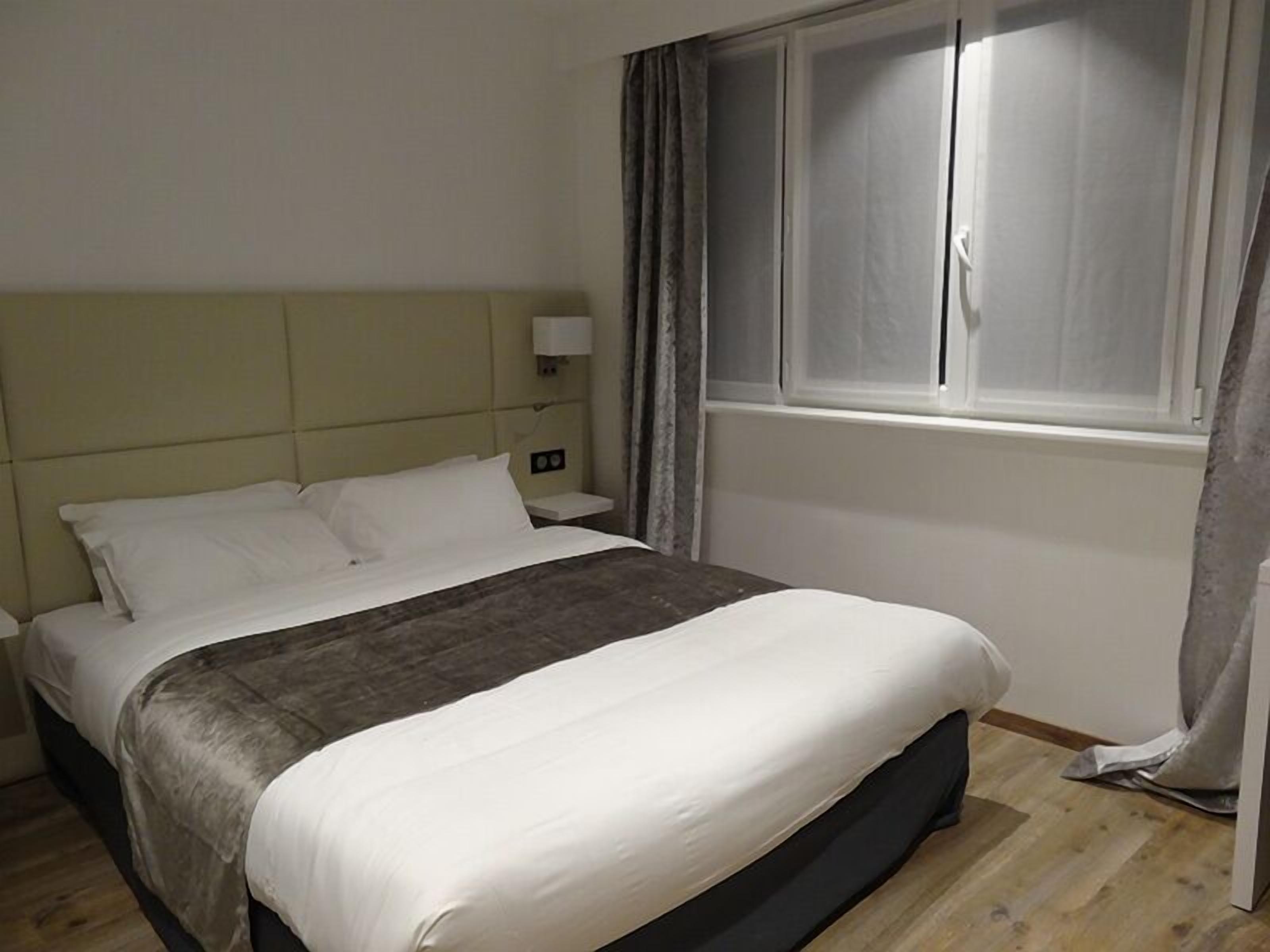 A neatly made bed with white and gray bedding, a bedside table with a lamp, and a window with curtains.