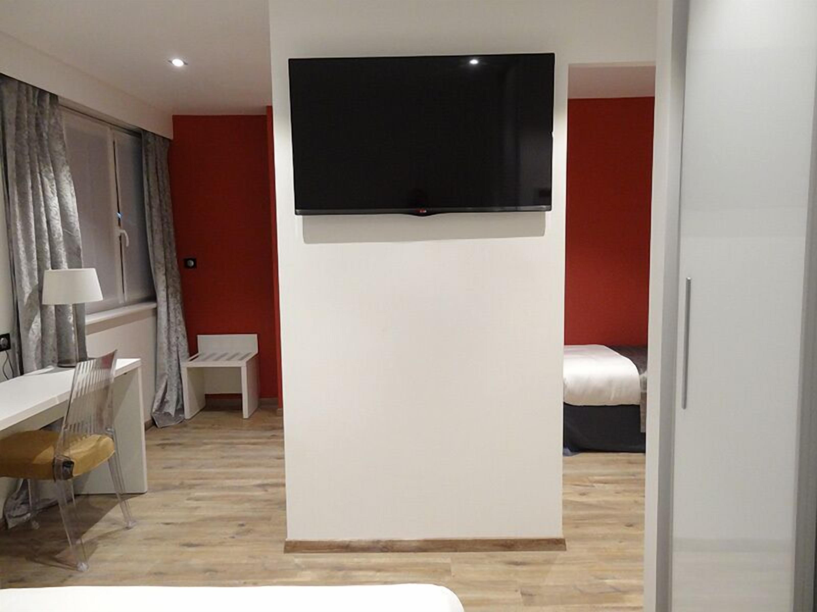 A modern hotel room with a flat-screen TV mounted on the wall, a desk with a chair, a bed, and a small table.