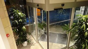 Rincón de un vestíbulo de hotel con puertas de cristal, plantas en macetas y suelo de baldosas.