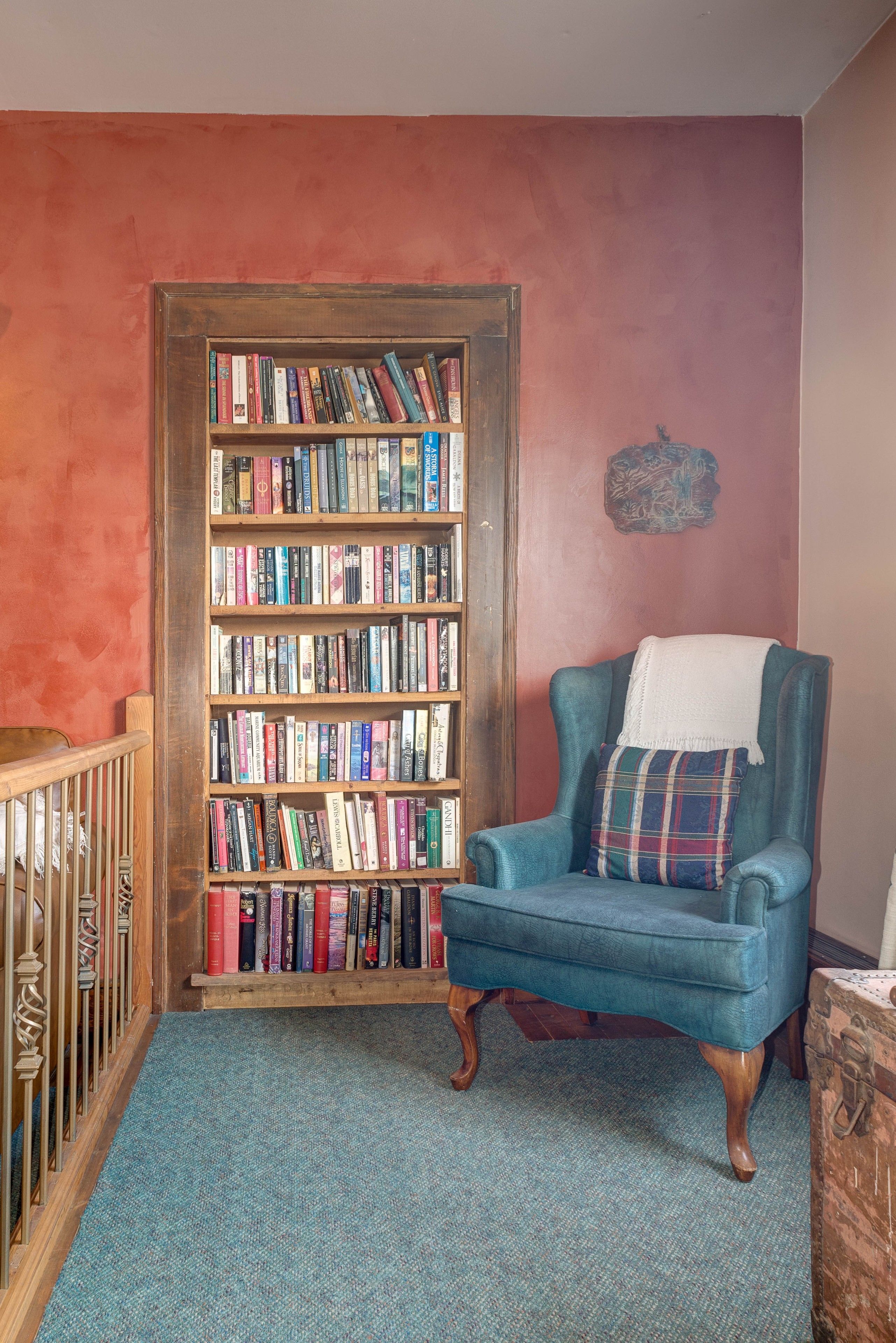 A room with a wooden bookshelf filled with books, a blue armchair with a plaid pillow, and a wooden crib.