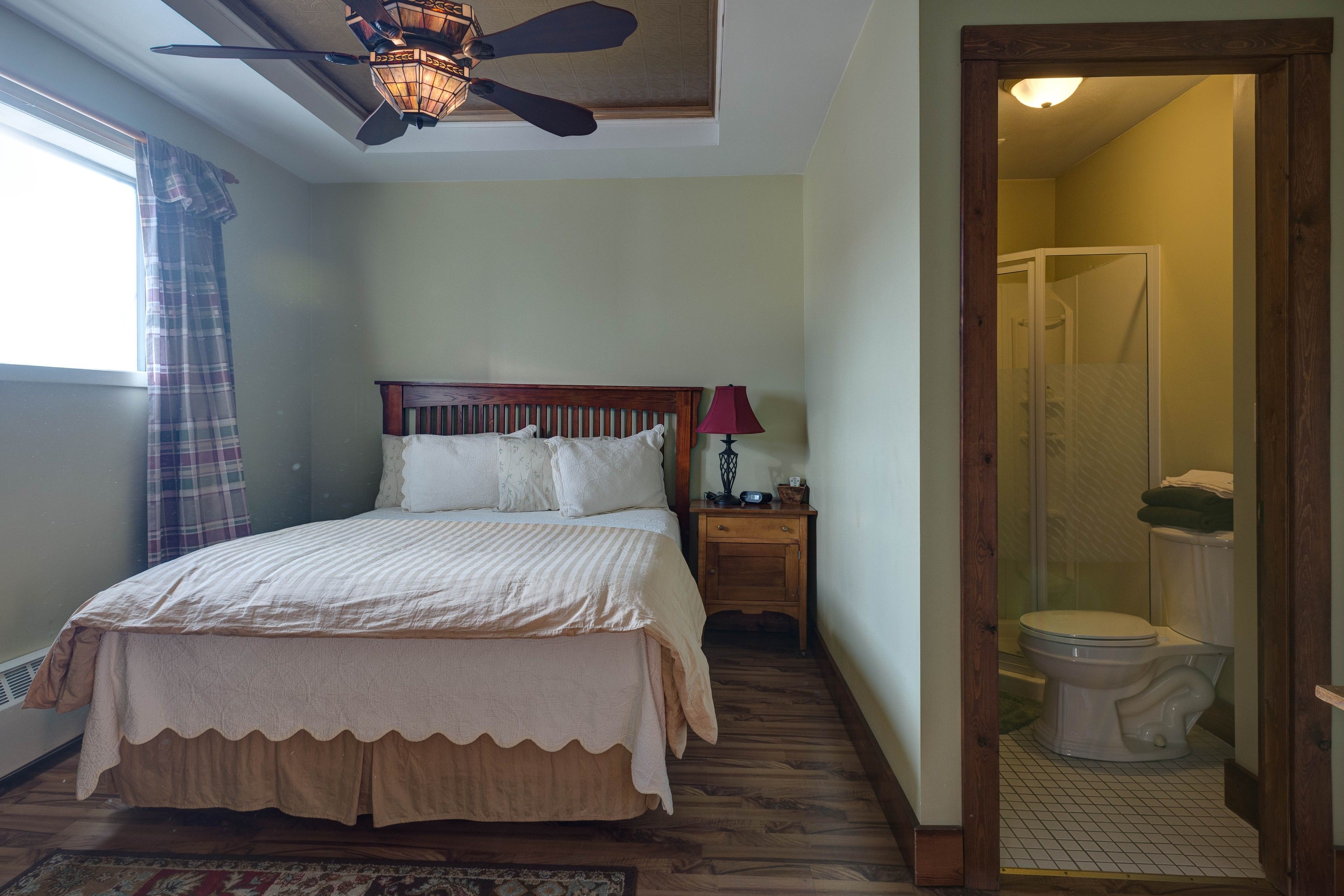 A bedroom with a bed, a nightstand, a lamp, a ceiling fan, and a view of the bathroom.