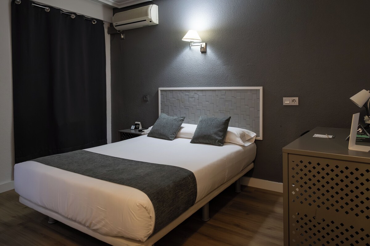 Una habitación de hotel con cama, mesita de noche, lámpara y aire acondicionado.