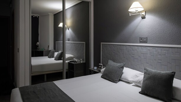 Habitación de hotel con cama, mesita de noche, lámpara, espejo y pared con diseño geométrico.